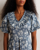 A woman wearing a dusty blue and white floral print dress with a ruffled Peter Pan collar and short puff sleeves, shown from the chest up against a neutral light background.