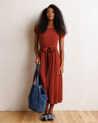 A woman stands against a textured off-white wall on a light wood floor, wearing a rust-colored short-sleeve midi dress with a tie waist and pockets, holding a blue tote bag.