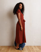A woman stands in profile wearing a rust-colored midi dress with puff sleeves and a matching belt, layered over blue jeans and black-and-white patterned slip-on shoes, against a textured off-white wall and light wood floor.