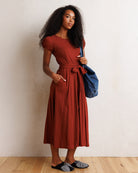 A woman stands against a textured off-white wall on a light wood floor, wearing a rust-colored short-sleeve midi dress with a tie waist and pockets, holding a blue tote bag.