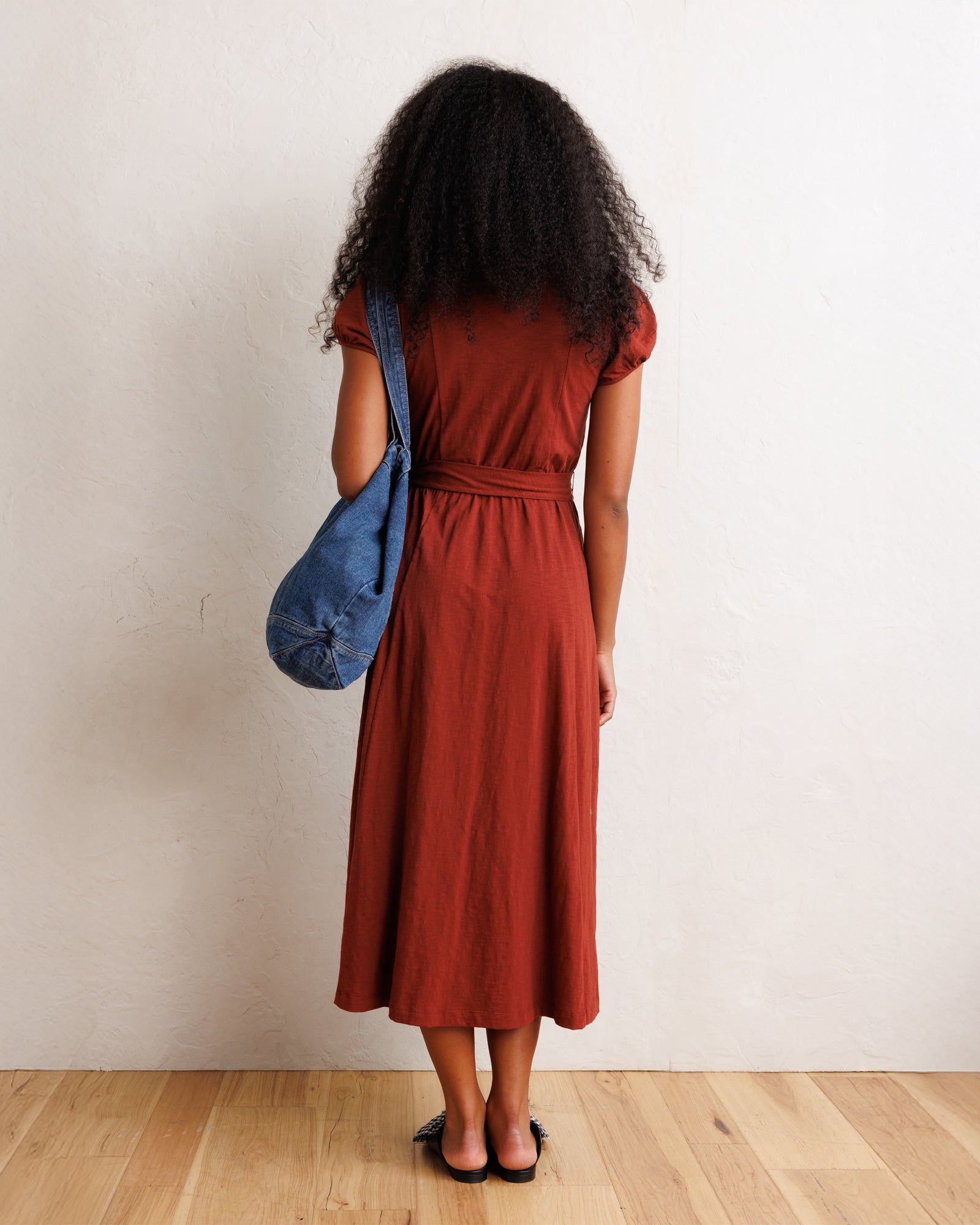 A woman with curly black hair is shown from behind wearing a rust-colored short-sleeve midi dress with a matching belt. She is carrying a blue denim shoulder bag and wearing black flat sandals, standing against a textured off-white wall on a light wood floor.