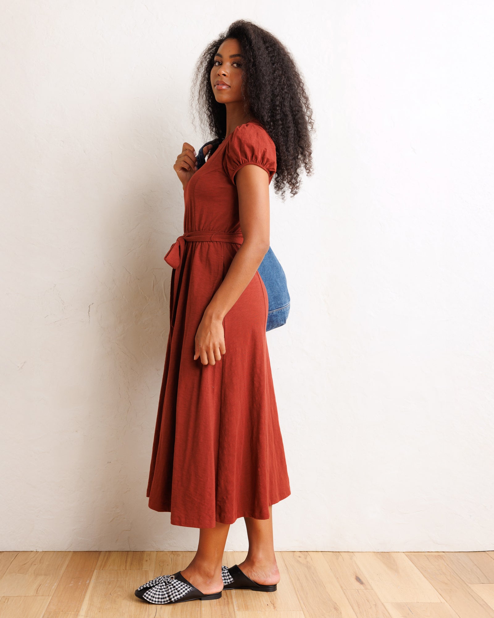 A woman stands in profile wearing a rust-red midi dress with short puff sleeves, a V-neckline, and a matching fabric tie belt at the waist. She is also wearing a blue shoulder bag and black-and-white patterned slip-on shoes, standing against a plain off-white wall on a light wood floor.