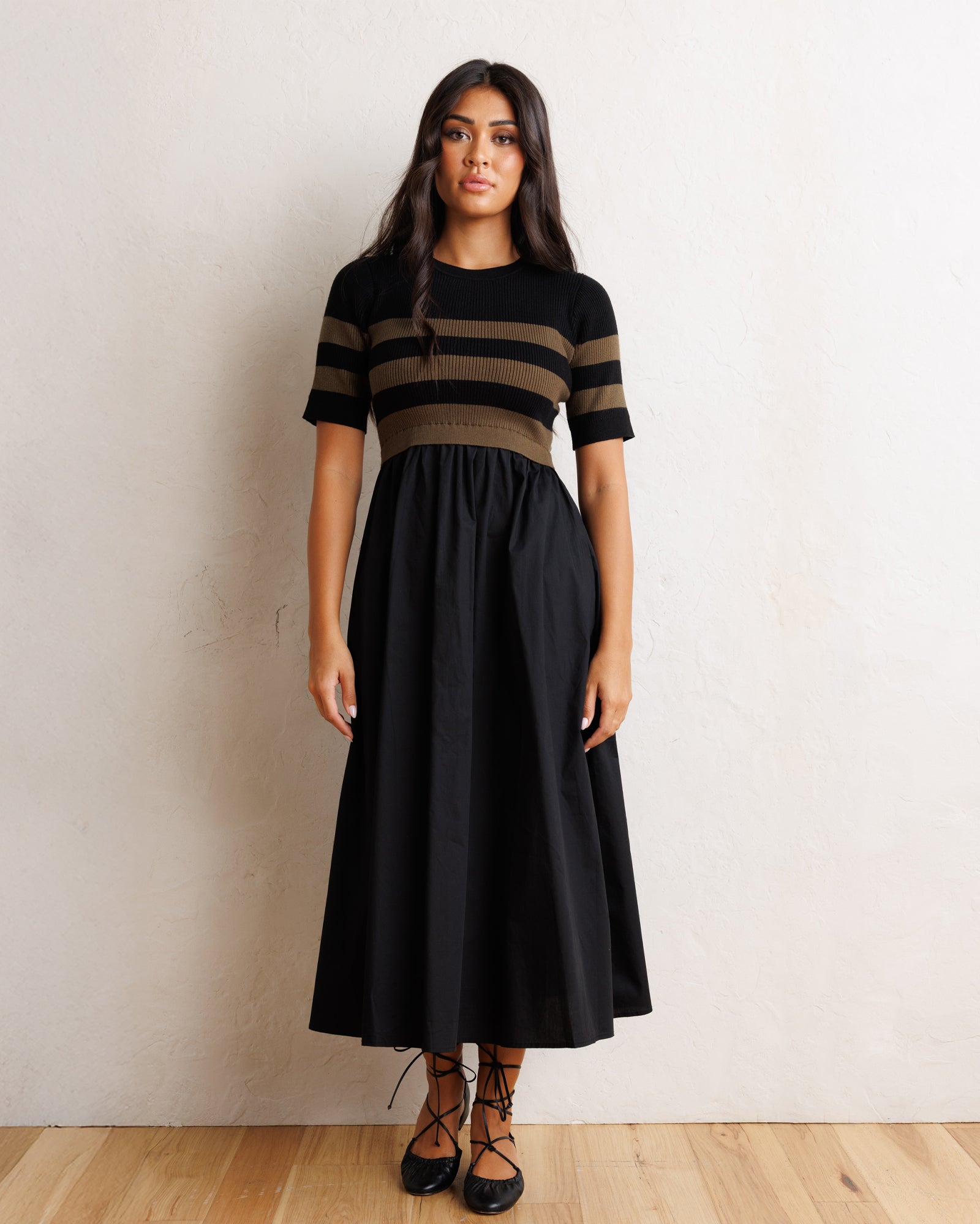A woman modeling a black midi dress with a short-sleeve top featuring horizontal black and brown stripes, standing against a textured off-white wall on a light wood floor.