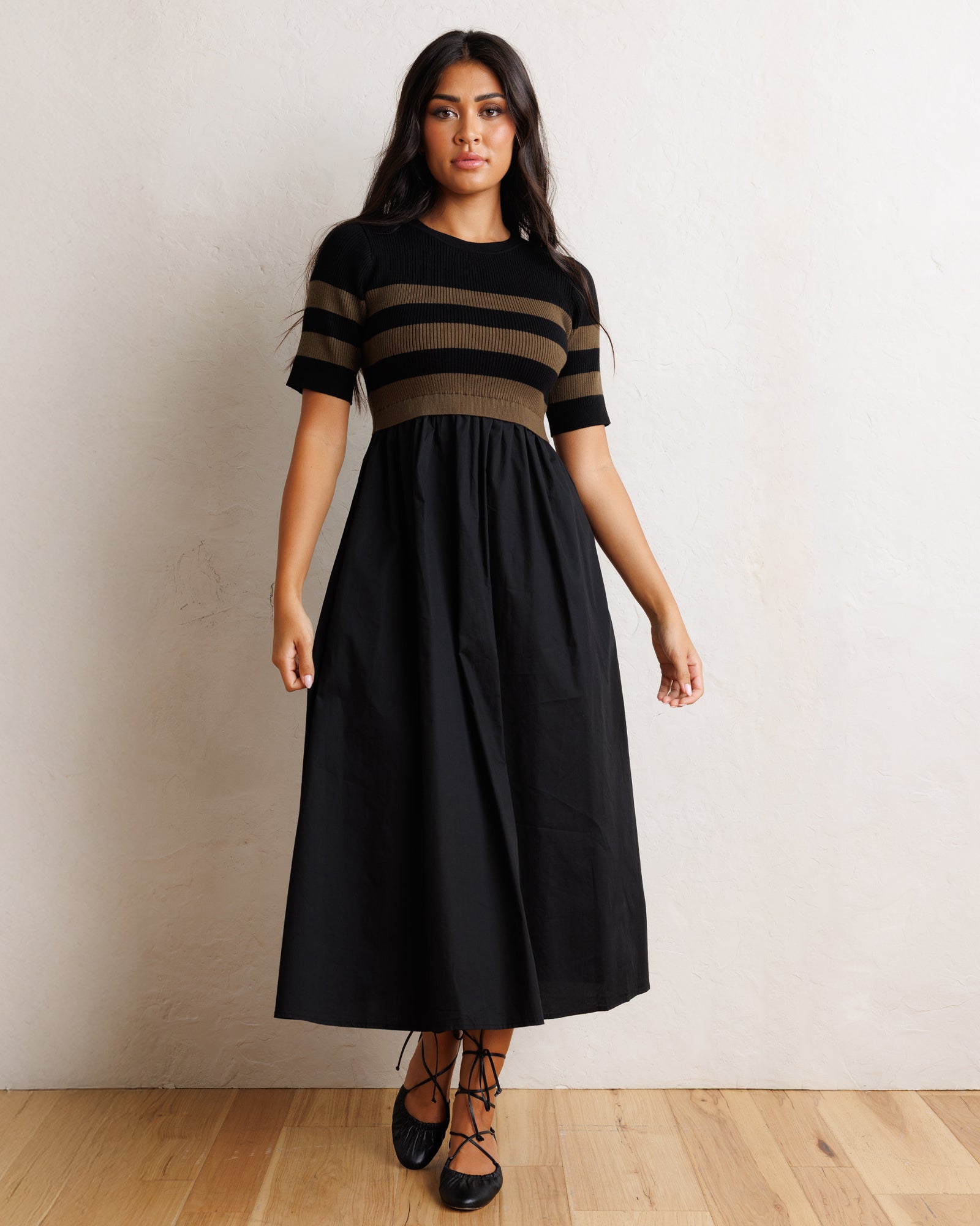A woman modeling a black midi dress with a short-sleeve top featuring horizontal black and brown stripes, paired with a gathered black skirt and black lace-up shoes, standing against a textured off-white wall and light wood floor.