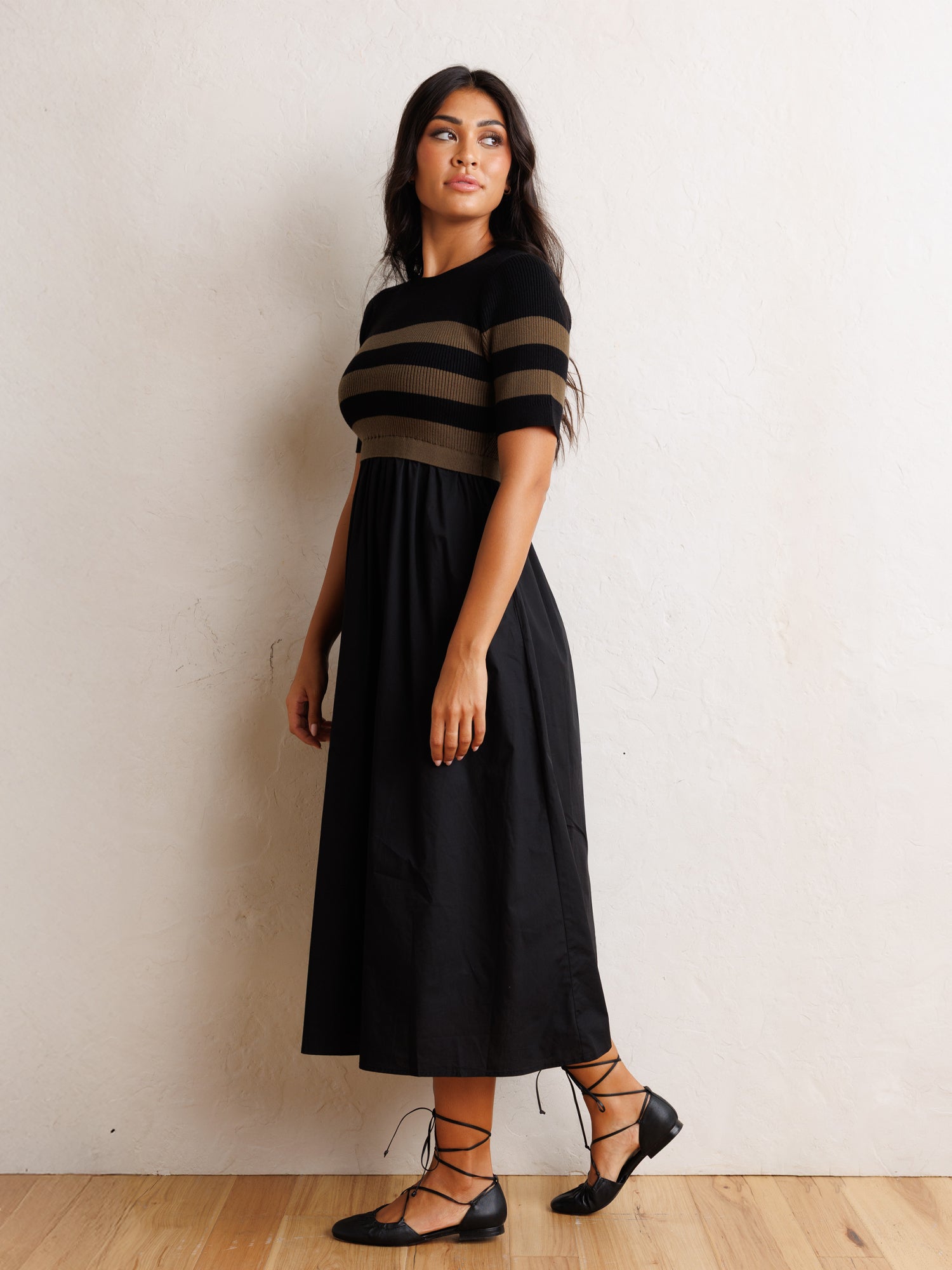 A woman modeling a black midi dress with a short-sleeve top featuring horizontal black and brown stripes, paired with black lace-up flats, standing against a textured off-white wall on a light wood floor.