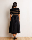 A woman is shown from behind wearing a black midi dress with brown horizontal stripes on the bodice and sleeves, paired with black lace-up heels, standing against a textured off-white wall on a light wood floor.