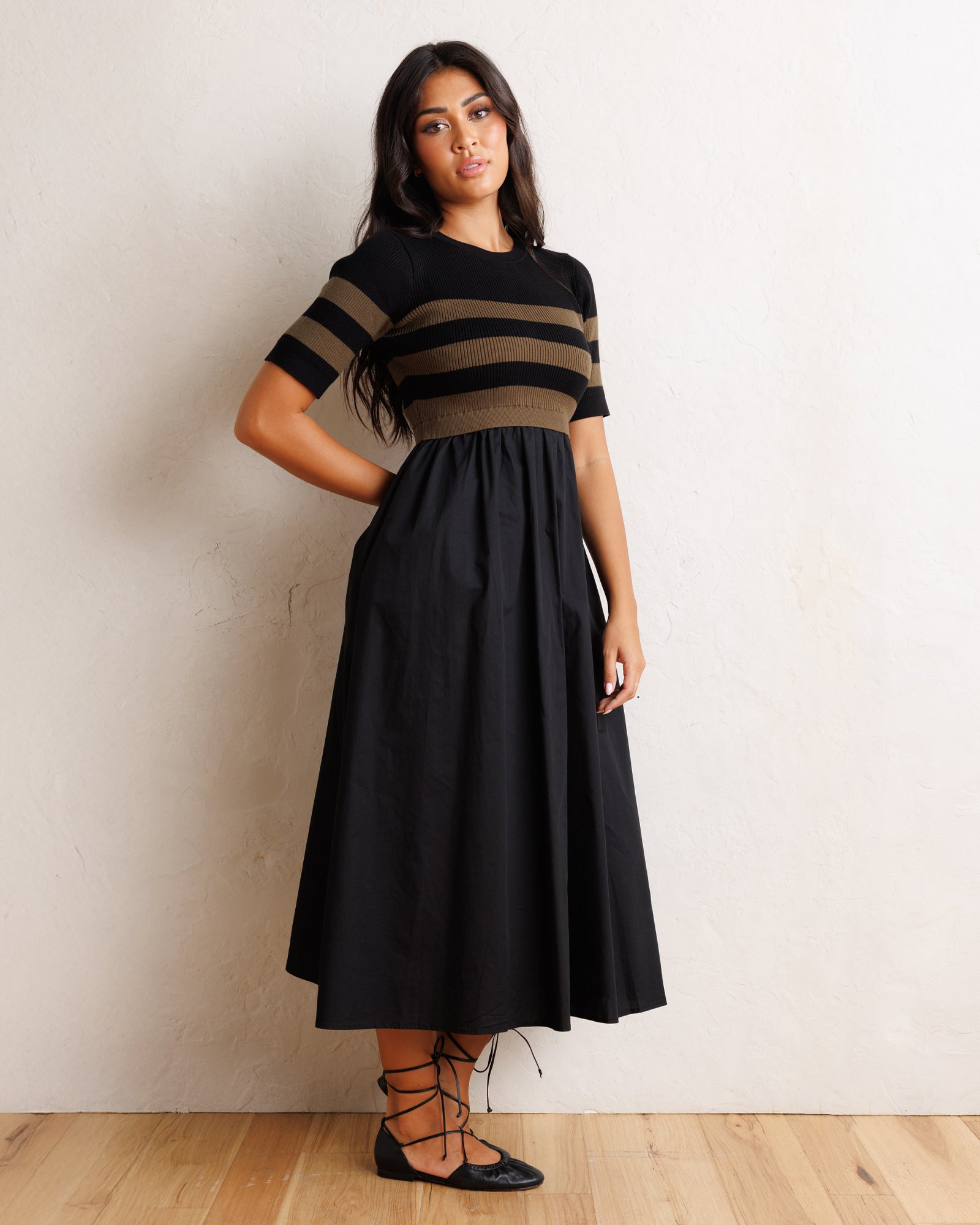 A woman modeling a midi dress with a black and olive green striped short-sleeve bodice and a solid black skirt, standing against a textured off-white wall on a light wood floor.