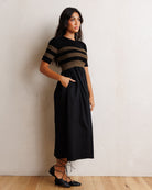 A woman modeling a black midi dress with a short-sleeve top featuring horizontal black and brown stripes, standing against a textured off-white wall on a light wood floor.
