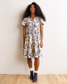 A woman modeling a short-sleeve tiered midi dress with a blue and yellow floral print on a cream background, paired with white socks and navy boots, standing against a white wall.