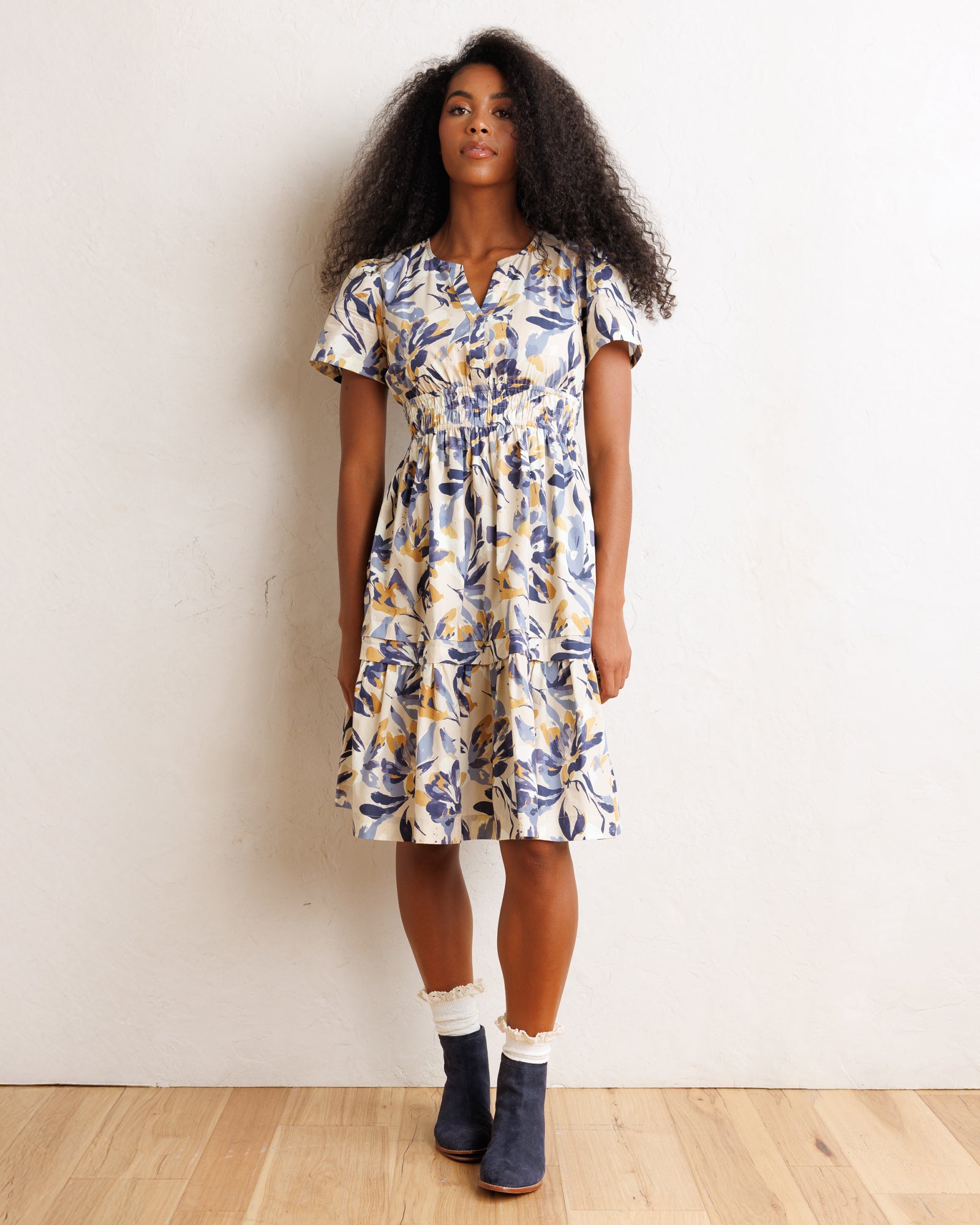 A woman modeling a short-sleeve tiered midi dress with a blue and yellow floral print on a cream background, paired with white socks and navy boots, standing against a white wall.