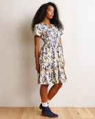 A woman modeling a short-sleeve midi dress with a blue, yellow, and white botanical leaf pattern, standing against a textured off-white wall on a light wood floor.