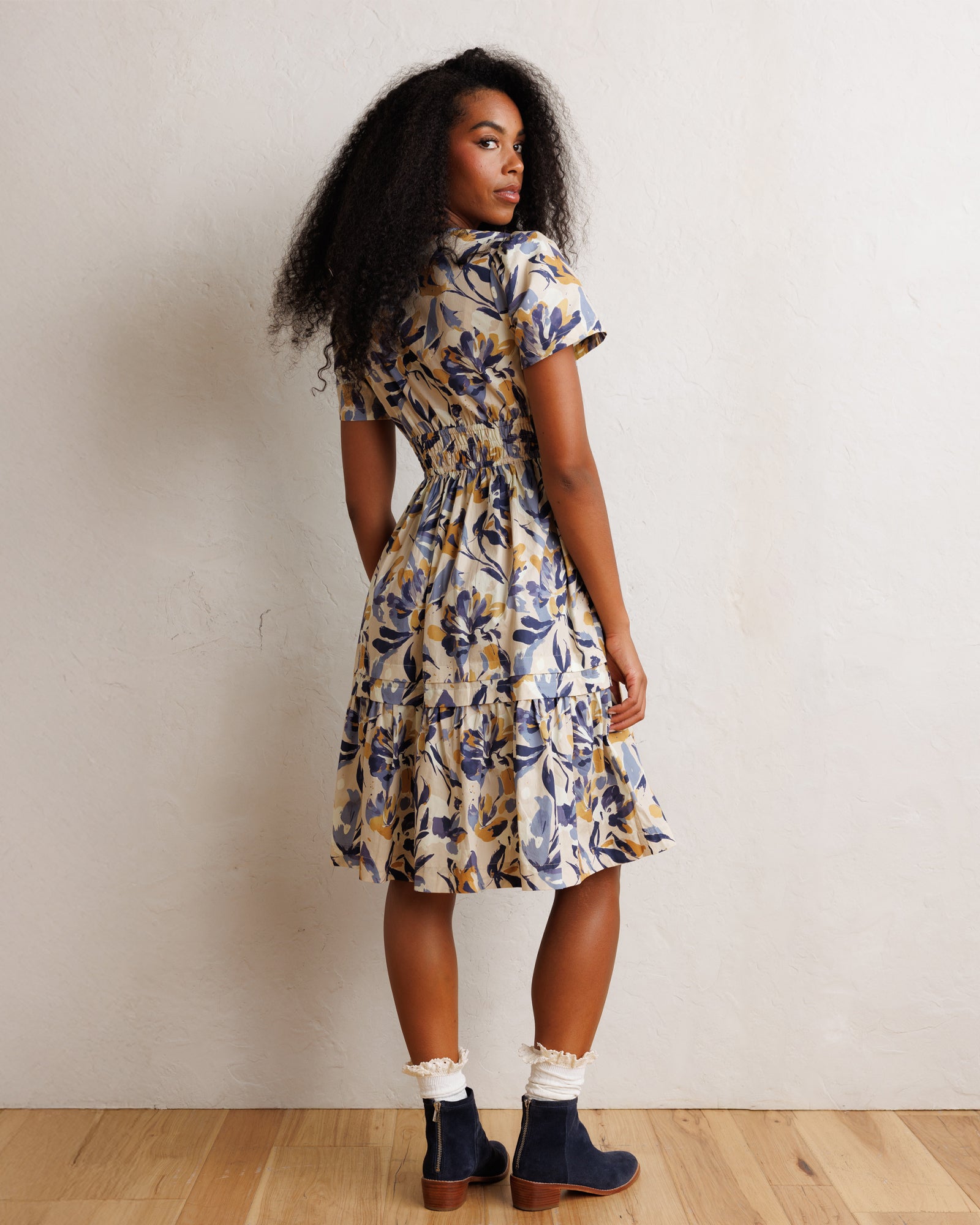A woman wearing a short-sleeve tiered midi dress with a blue and yellow floral pattern on a cream background, standing against a textured off-white wall on a light wood floor.