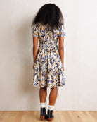A woman viewed from behind wearing a short-sleeve tiered midi dress with a blue, yellow, and white botanical pattern, paired with white ruffled socks and navy ankle boots, standing against a textured off-white wall on a light wood floor.