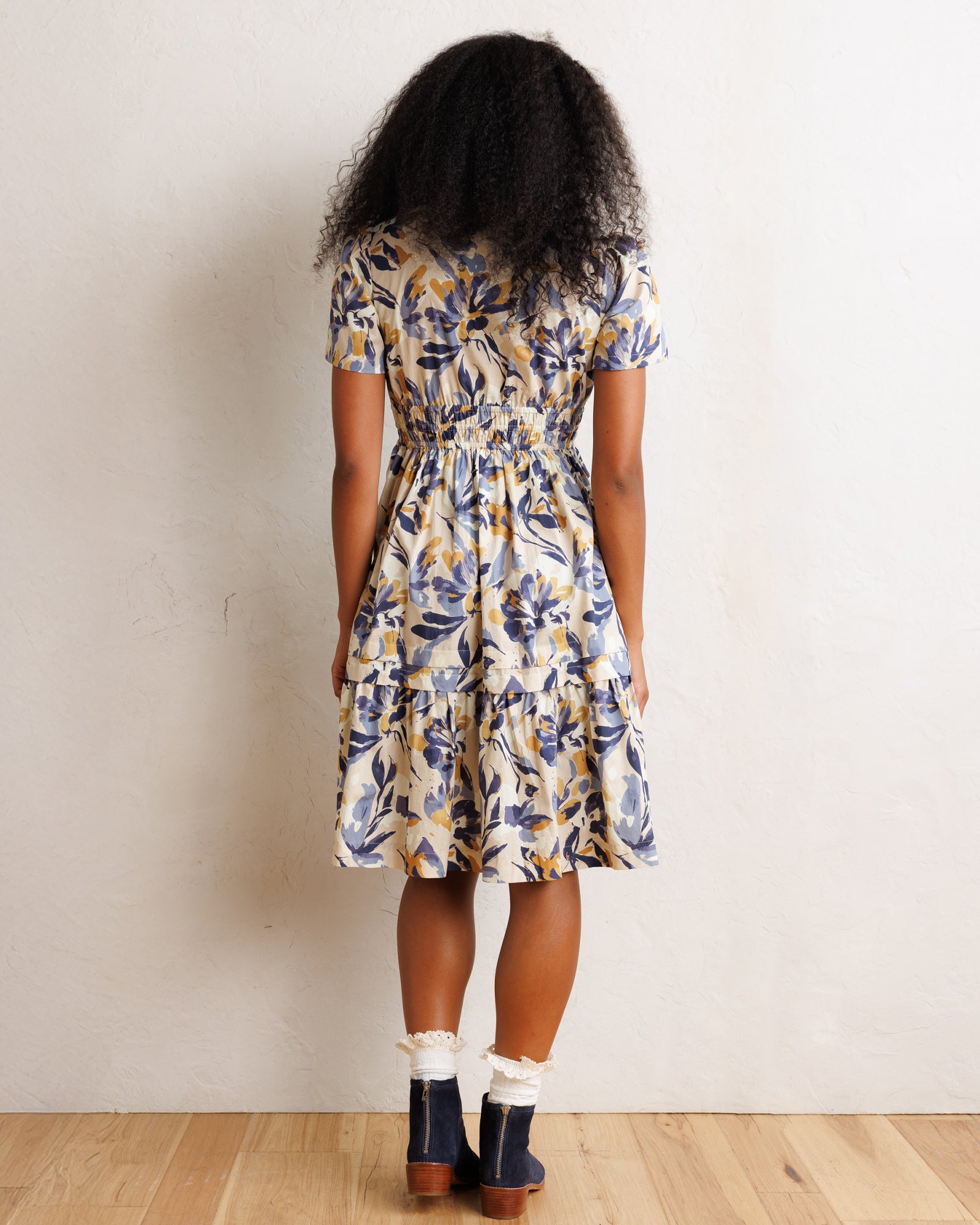 A woman viewed from behind wearing a short-sleeve tiered midi dress with a blue, yellow, and white botanical pattern, paired with white ruffled socks and navy ankle boots, standing against a textured off-white wall on a light wood floor.