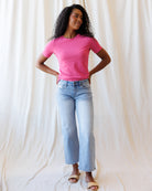 A woman wearing a pink short-sleeve crewneck t-shirt and light blue jeans, standing against a fabric backdrop.