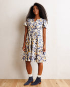 A woman modeling a short-sleeve tiered midi dress with a blue and yellow botanical leaf pattern on a cream background, paired with navy ankle boots and white socks, standing against a plain off-white wall.