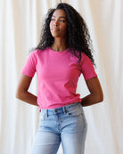 A woman wearing a pink short-sleeve crewneck t-shirt and light blue jeans, standing against a fabric backdrop.