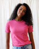 A woman with long, curly black hair wearing a bright pink short-sleeve crewneck t-shirt and light wash blue jeans, standing against a white curtain background.