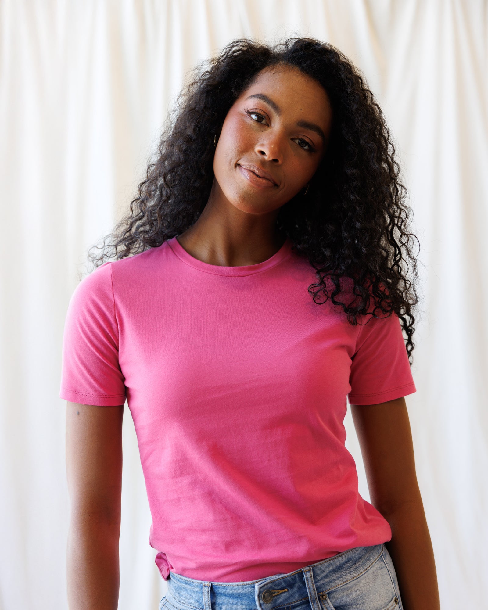 A woman with long, curly black hair wearing a bright pink short-sleeve crewneck t-shirt and light wash blue jeans, standing against a white curtain background.