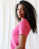 A woman wearing a pink short-sleeve crewneck t-shirt and light blue jeans, standing against a fabric backdrop.