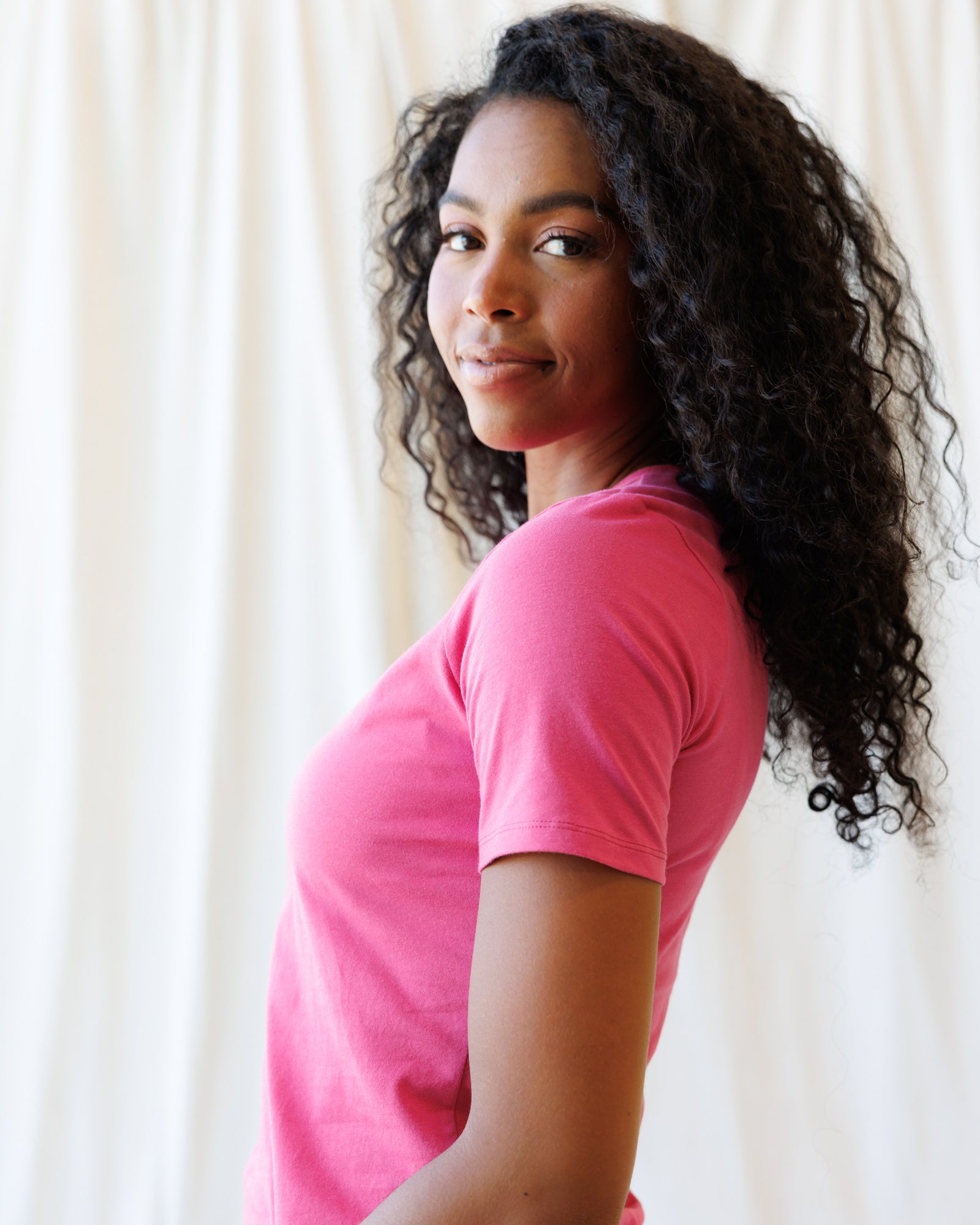 A woman wearing a pink short-sleeve crewneck t-shirt and light blue jeans, standing against a fabric backdrop.