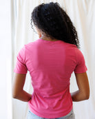A woman wearing a pink short-sleeve crewneck t-shirt and light blue jeans, standing against a fabric backdrop.
