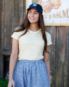 Woman wearing a yellow top and blue checkered skirt with a navy cap, standing in front of a wooden wall.