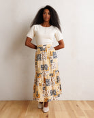 A woman stands against a textured off-white wall on a light wood floor, wearing a cream ribbed short-sleeve top and a tiered maxi skirt with a yellow, black, and white patchwork pattern.