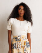 A woman with long, curly black hair stands against a textured off-white wall, wearing a cream ribbed short-sleeve henley top and a yellow, navy, and white patterned skirt.
