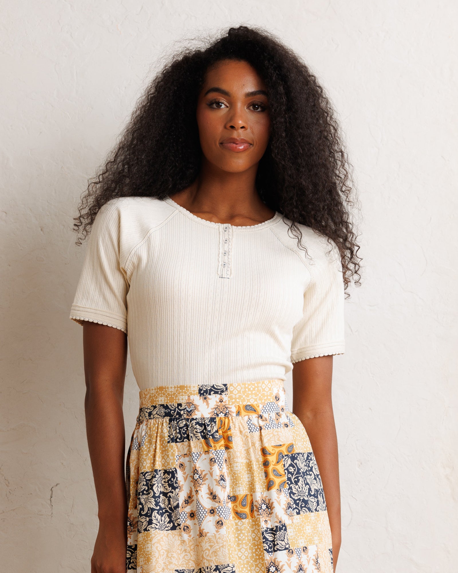 A woman with long, curly black hair stands against a textured off-white wall, wearing a cream ribbed short-sleeve henley top and a yellow, navy, and white patterned skirt.