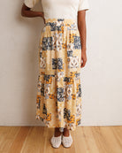 A model wearing a tiered, long maxi skirt with a patchwork print of yellow, black, and white patterns, paired with a cream top and white crochet flats, standing against a textured off-white wall and light wood floor.