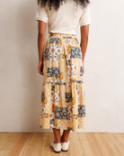 A person shown from the back wearing a cream ribbed short-sleeve top and a tiered midi skirt with a patchwork-style print in beige, mustard yellow, navy, gray, and white. The person is standing on a light wood floor against a textured off-white wall.