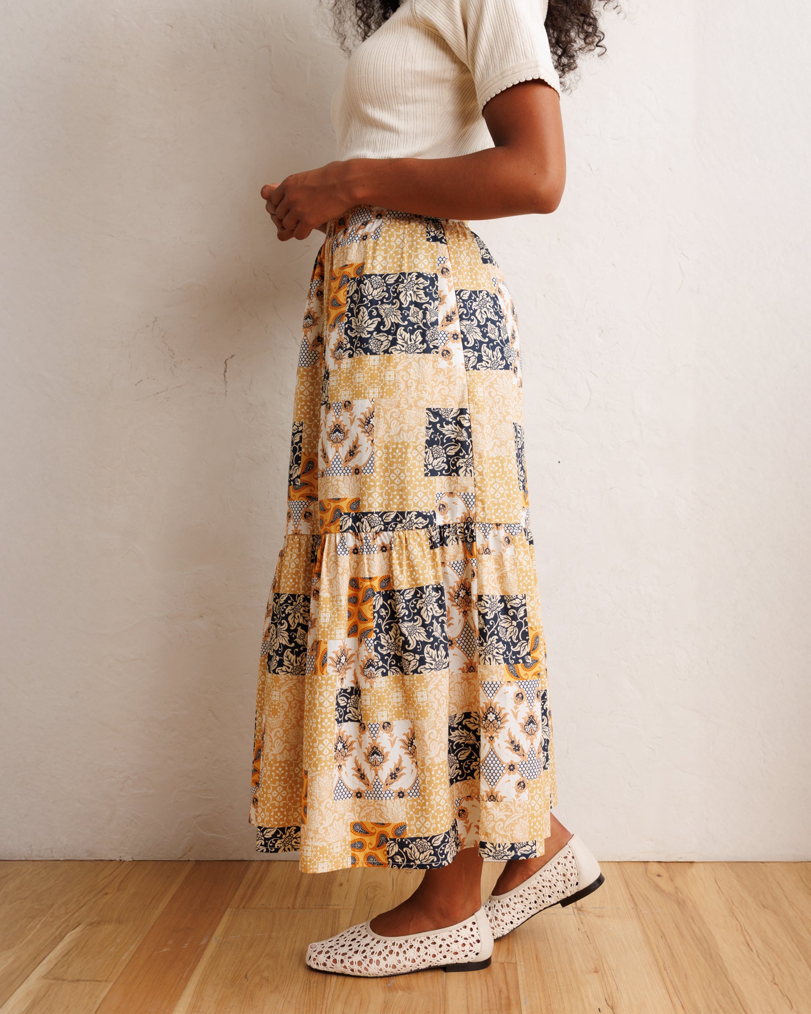 A woman wearing a tiered maxi skirt with a patchwork print in yellow, navy, and white, paired with a cream short-sleeve top and white patterned flats, standing against a textured off-white wall on a light wood floor.