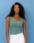 A woman with long, curly black hair wearing a fitted sage green scoop-neck tank top and white jeans, standing against a solid blue background.