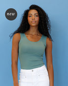 A woman with long curly black hair wearing a sage green ribbed scoop-neck tank top and white pants, standing against a solid blue background with a black 'new' badge overlay in the top left.