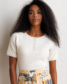 A woman with long, curly black hair stands against a textured off-white wall, wearing a cream ribbed short-sleeve henley top and a yellow, navy, and white patterned skirt.