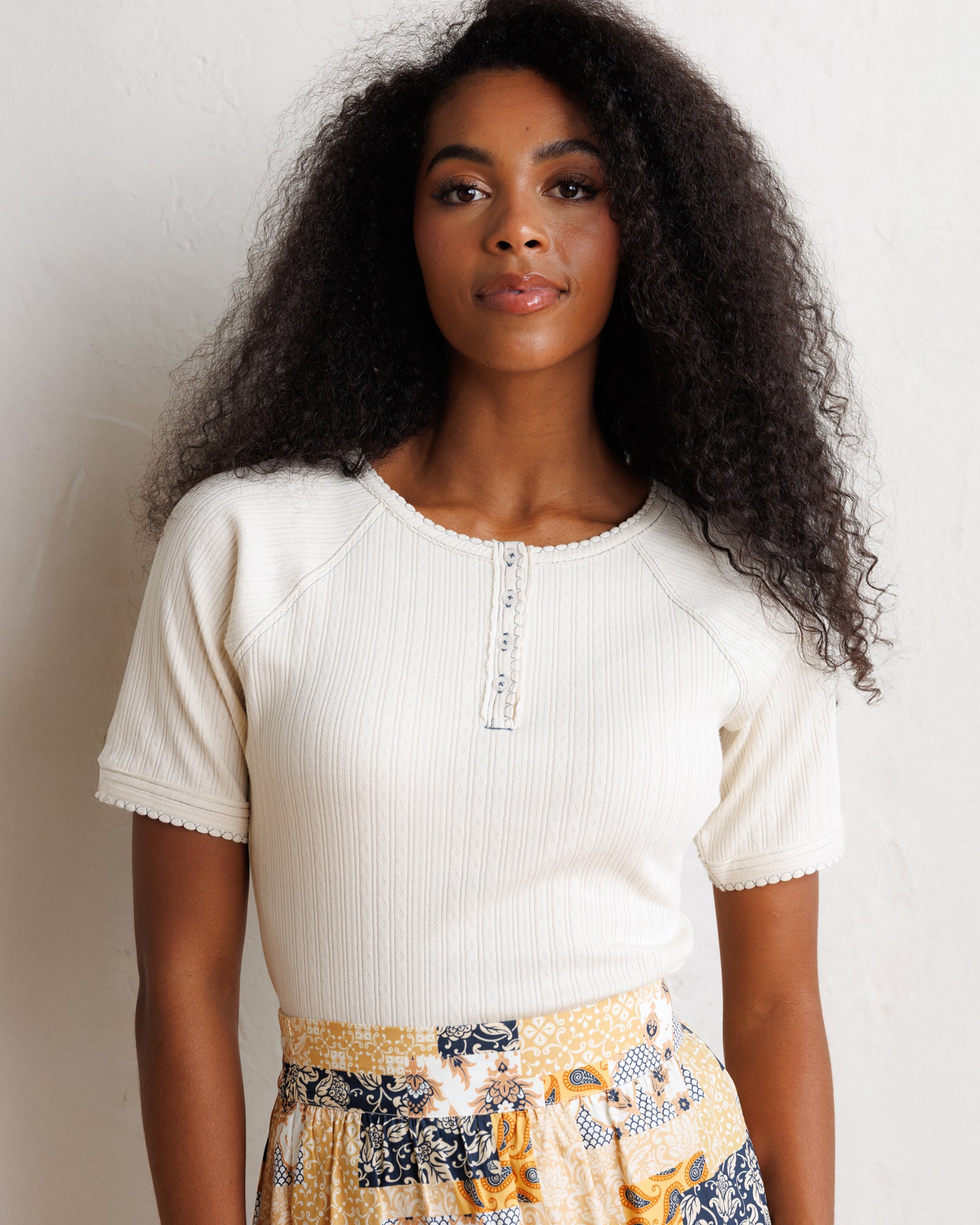 A woman with long, curly black hair stands against a textured off-white wall, wearing a cream ribbed short-sleeve henley top and a yellow, navy, and white patterned skirt.