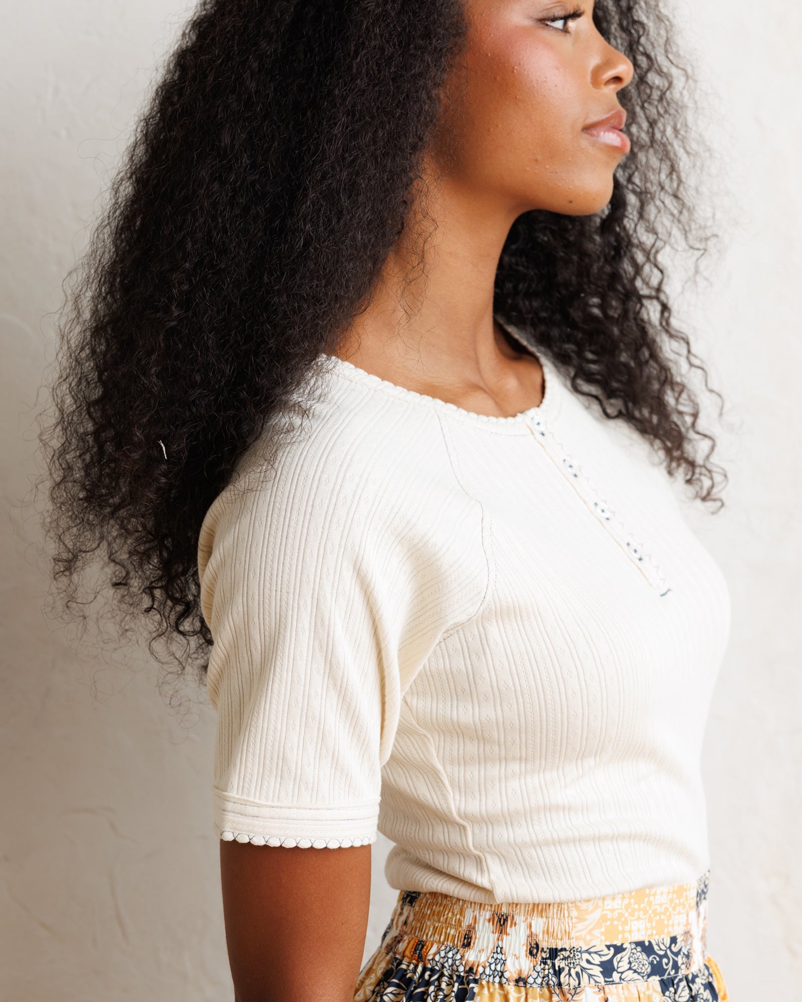 A woman shown in profile wearing a ribbed short-sleeve cream top with a scalloped hem and a small decorative embellishment along the neckline, paired with a patterned skirt against a plain light wall.