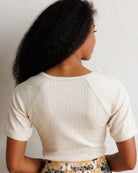 A woman shown from behind wearing a cream-colored ribbed short-sleeve crop top with scalloped edges, paired with a patterned skirt, against a textured off-white wall.