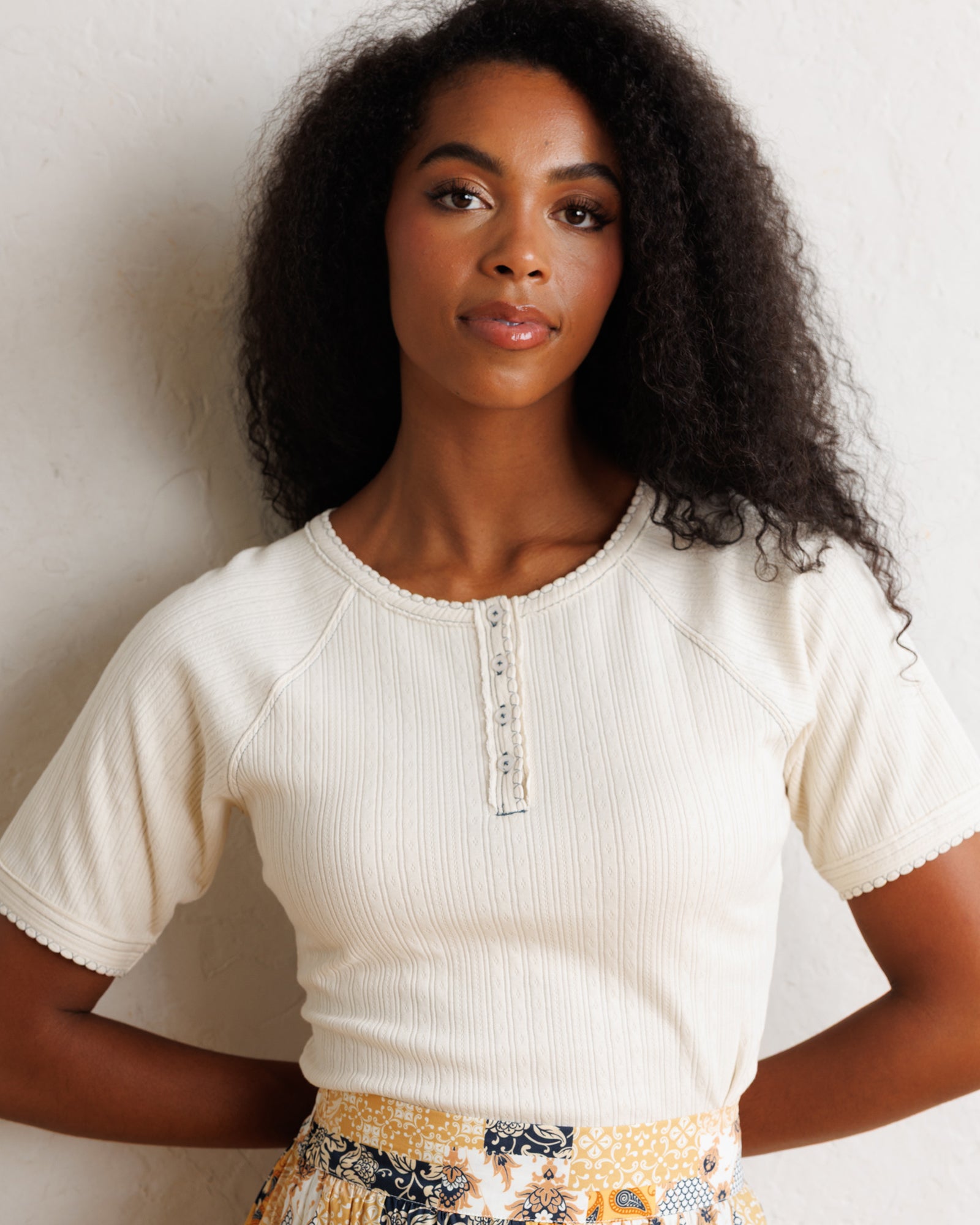A woman with curly dark hair wearing a ribbed cream short-sleeve henley top and a yellow patterned skirt, posing against a textured off-white wall.