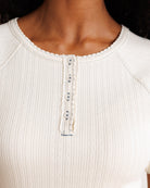 Close-up of a cream ribbed knit top worn by a model, featuring a scalloped neckline and a button placket with blue embroidered accents.
