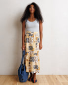 A woman stands against a white wall on a light wood floor, wearing a light gray tank top and a long cream maxi skirt with a yellow, black, and gray abstract print. She is holding a large blue denim tote bag.