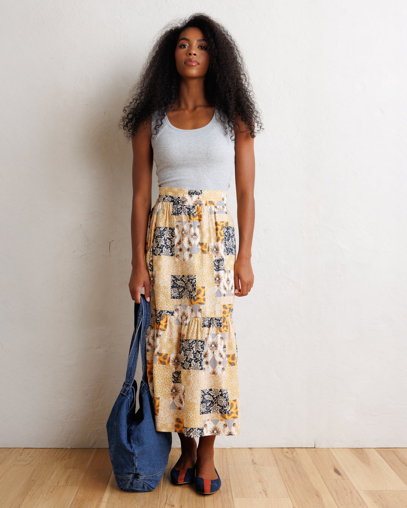 A woman stands against a white wall on a light wood floor, wearing a light gray tank top and a long cream maxi skirt with a yellow, black, and gray abstract print. She is holding a large blue denim tote bag.