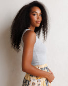 A woman with long, curly black hair is shown from the waist up, wearing a light gray sleeveless top and a patterned skirt with yellow, black, and white tones. She is posing against a textured off-white wall.