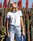 Woman standing in front of red floral plants wearing a white t-shirt with blue trim and light blue jeans.