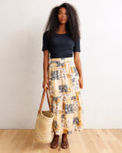 A woman stands in a room with a white wall and light wood floor, wearing a navy blue short-sleeve top and a long cream skirt with a blue and yellow abstract pattern. She holds a large woven straw tote bag.