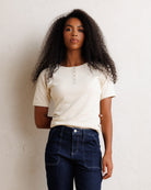 A woman with long, curly dark hair stands against a textured off-white wall, wearing a cream-colored short-sleeve ribbed top with a decorative button placket and dark blue jeans.