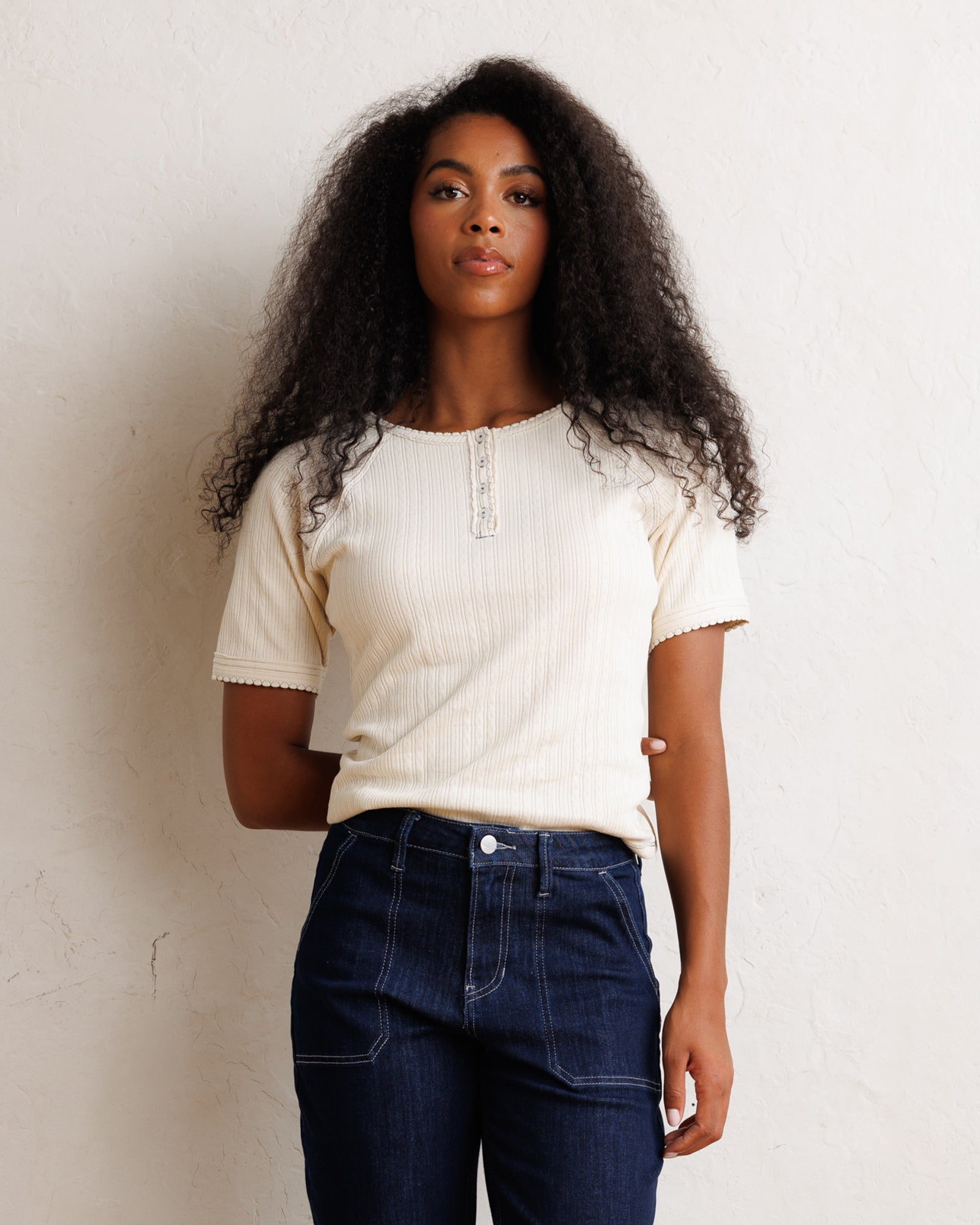 A woman with long, curly dark hair stands against a textured off-white wall, wearing a cream-colored short-sleeve ribbed top with a decorative button placket and dark blue jeans.