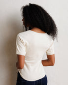 A woman with curly black hair is shown from the back wearing a cream-colored ribbed short-sleeve top and dark blue jeans, standing against a textured off-white wall.