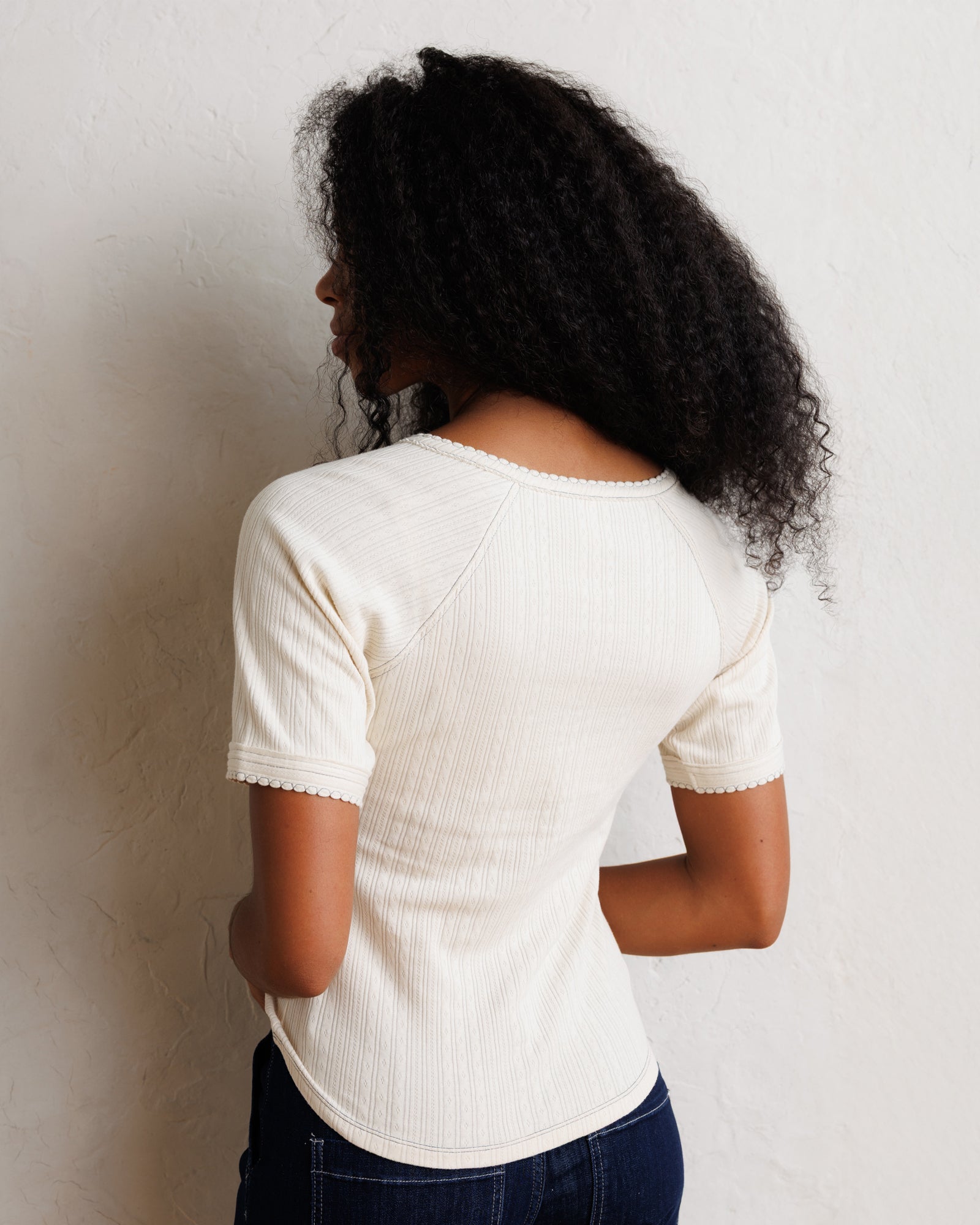 A woman with curly black hair is shown from the back wearing a cream-colored ribbed short-sleeve top and dark blue jeans, standing against a textured off-white wall.